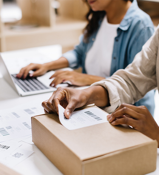 Person beschriftet ein Versandpaket und tippt gleichzeitig auf einem Laptop – barrierefreie Bestellprozesse und Retourenabwicklung für Online-Shops nach BFSG