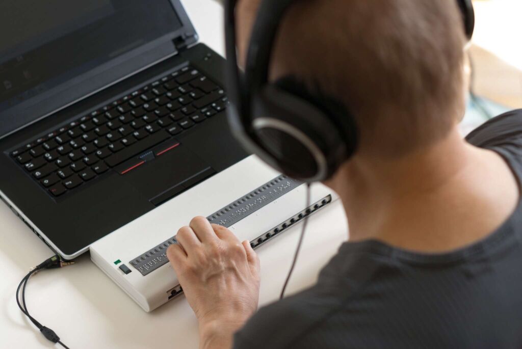 Person mit Kopfhörern bedient eine Braillezeile vor einem Laptop – Was ist ein Screenreader? Funktion, Bedeutung und Auswirkung auf die Website-Barrierefreiheit nach WCAG 2.2 und BFSG