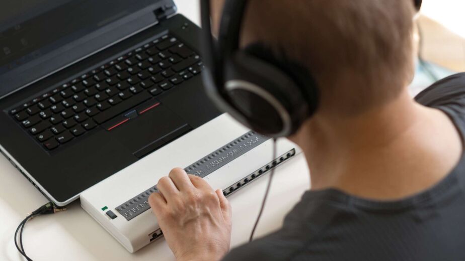 Person mit Kopfhörern bedient eine Braillezeile vor einem Laptop – Was ist ein Screenreader? Funktion, Bedeutung und Auswirkung auf die Website-Barrierefreiheit nach WCAG 2.2 und BFSG