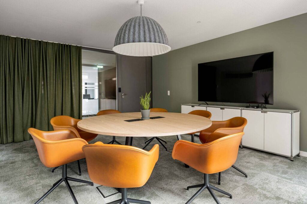 Moderner Konferenzraum mit rundem Holztisch und orangen Designerstühlen – Beratungsstandort der IFDB GmbH für digitale Barrierefreiheit und BFSG-Compliance in München