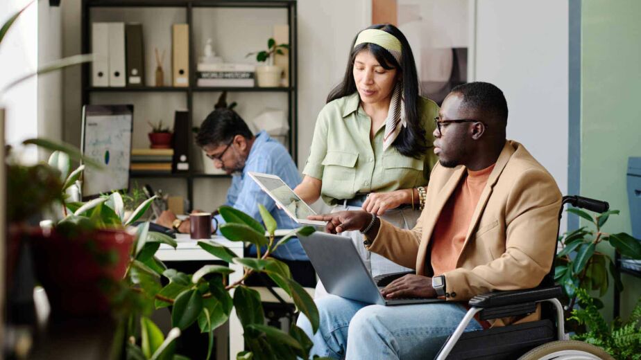 Mann im Rollstuhl arbeitet gemeinsam mit einer Kollegin am Laptop in einem modernen Büro – Barrierefreiheitsstärkungsgesetz: Pflichten, Fristen und Bußgelder für Unternehmen nach BFSG und EAA im Überblick