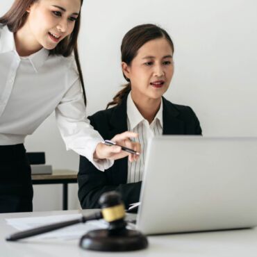 Zwei Frauen arbeiten gemeinsam am Laptop, im Vordergrund Richterhammer und Justitia-Waage – barrierefreie Formulare gestalten: Labels, Fehlermeldungen und Validierung nach WCAG 2.2 und BFSG