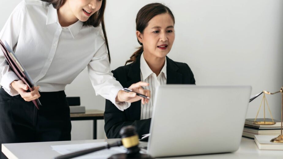 Zwei Frauen arbeiten gemeinsam am Laptop, im Vordergrund Richterhammer und Justitia-Waage – barrierefreie Formulare gestalten: Labels, Fehlermeldungen und Validierung nach WCAG 2.2 und BFSG