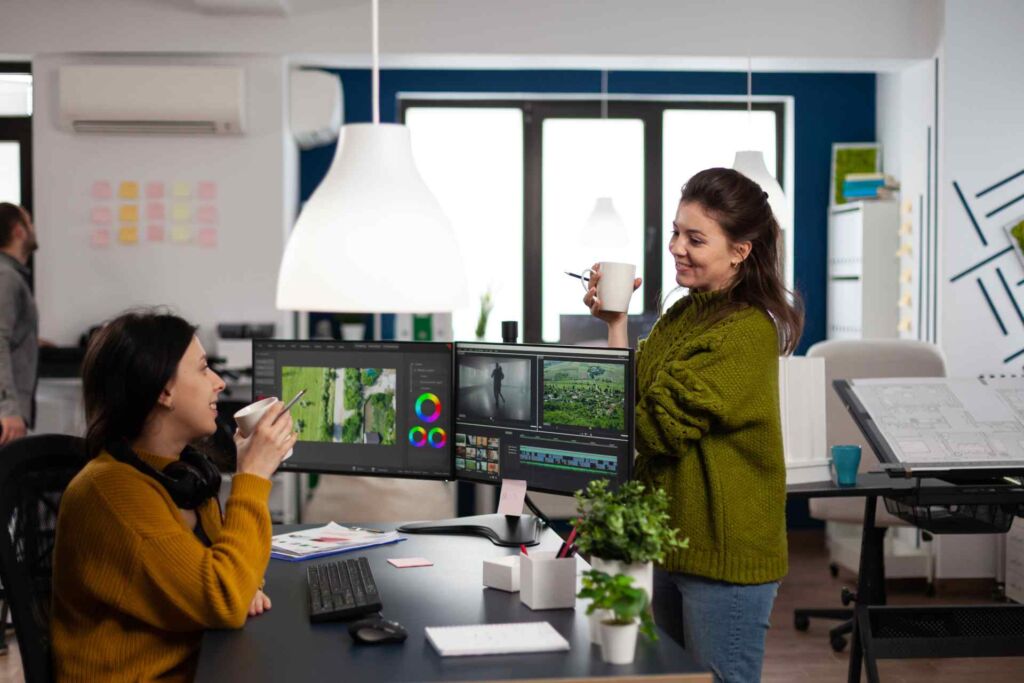 Zwei Designerinnen im lockeren Austausch vor einem Multi-Monitor-Setup mit Grafikbearbeitungssoftware – barrierefreies Webdesign: Prinzipien für kontrastreiche, tastaturzugängliche und screenreaderkompatible Websites nach WCAG 2.2