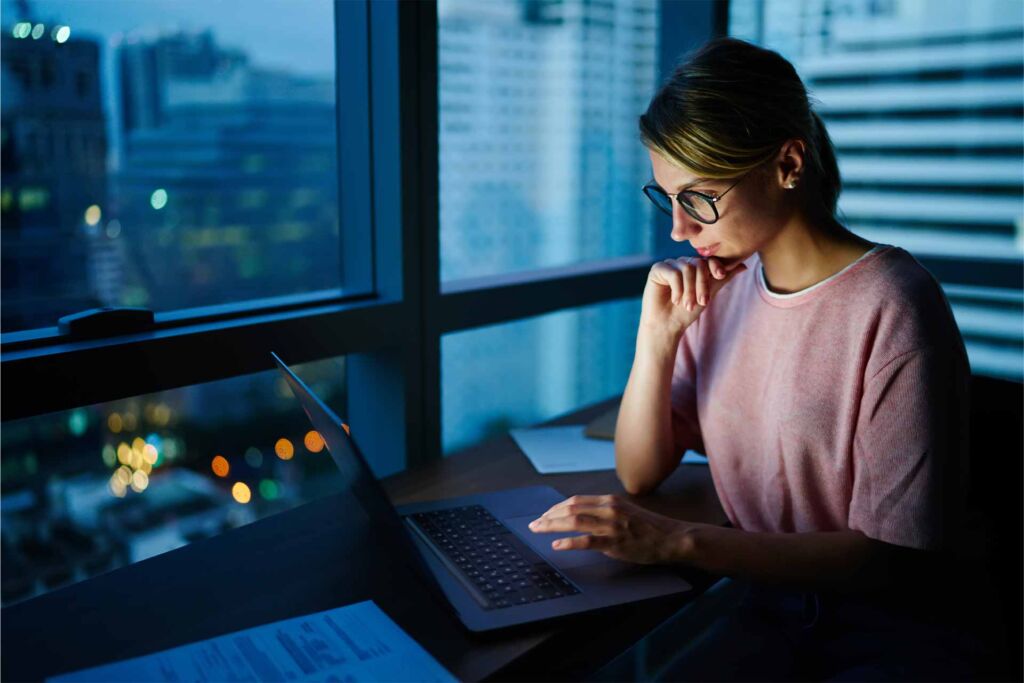 Frau mit Brille arbeitet konzentriert am Laptop vor nächtlicher Stadtkulisse – semantisches HTML als technisches Fundament barrierefreier Websites nach WCAG 2.2 und B