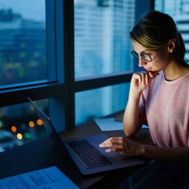 Frau mit Brille arbeitet konzentriert am Laptop vor nächtlicher Stadtkulisse – semantisches HTML als technisches Fundament barrierefreier Websites nach WCAG 2.2 und B