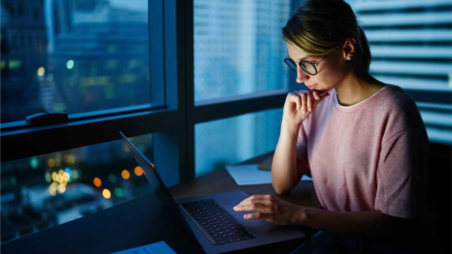 Frau mit Brille arbeitet konzentriert am Laptop vor nächtlicher Stadtkulisse – semantisches HTML als technisches Fundament barrierefreier Websites nach WCAG 2.2 und B