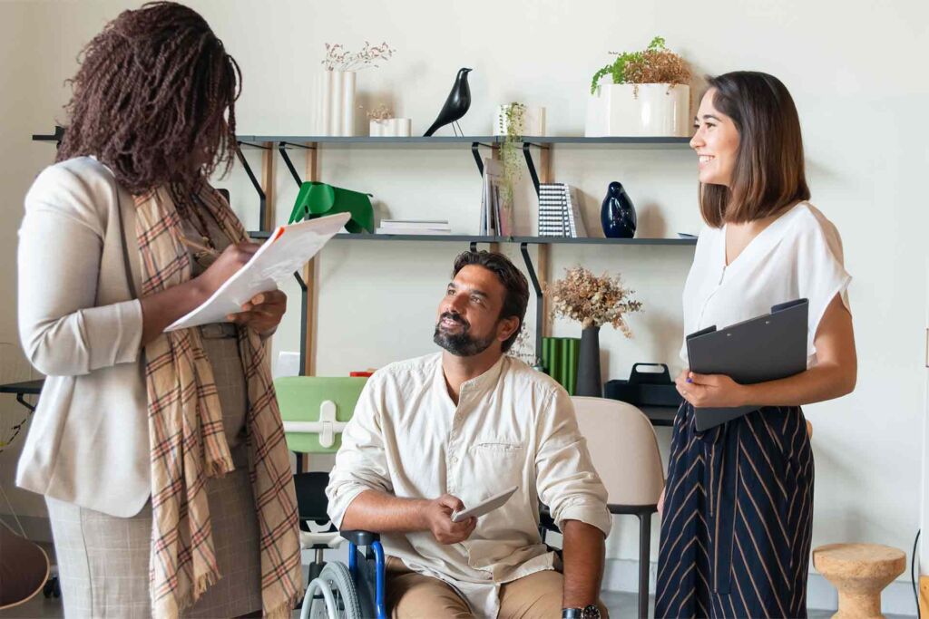 Drei Kollegen im Büro im Austausch, einer im Rollstuhl – barrierefreier Bewerbungsprozess für Menschen mit Beeinträchtigung