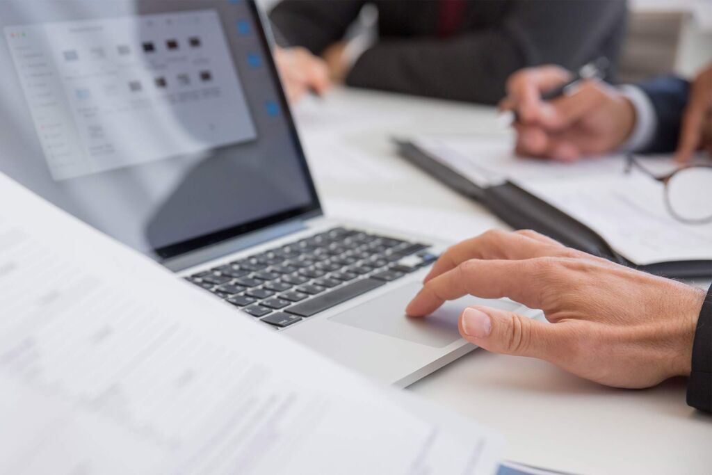 Hand auf Laptop-Trackpad neben Dokumenten im Meeting – PDF barrierefrei erstellen nach WCAG und PDF/UA