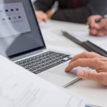 Hand auf Laptop-Trackpad neben Dokumenten im Meeting – PDF barrierefrei erstellen nach WCAG und PDF/UA
