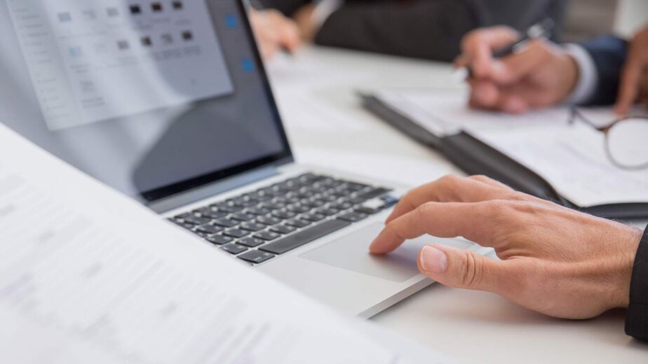 Hand auf Laptop-Trackpad neben Dokumenten im Meeting – PDF barrierefrei erstellen nach WCAG und PDF/UA